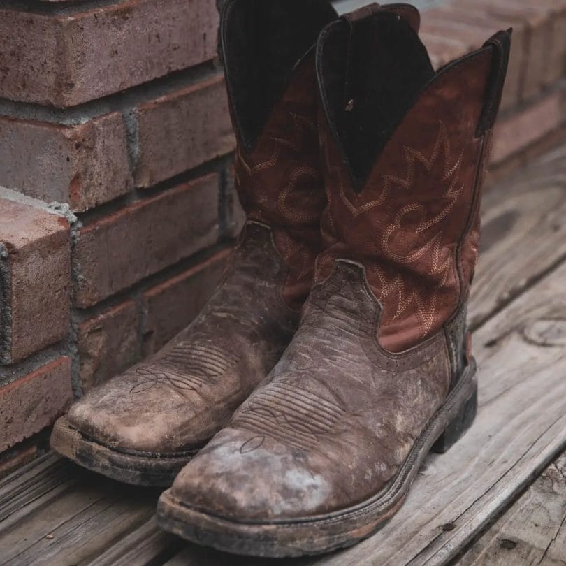 Pair of old cowboy boots sitting on a wooden bench next to red brick wall