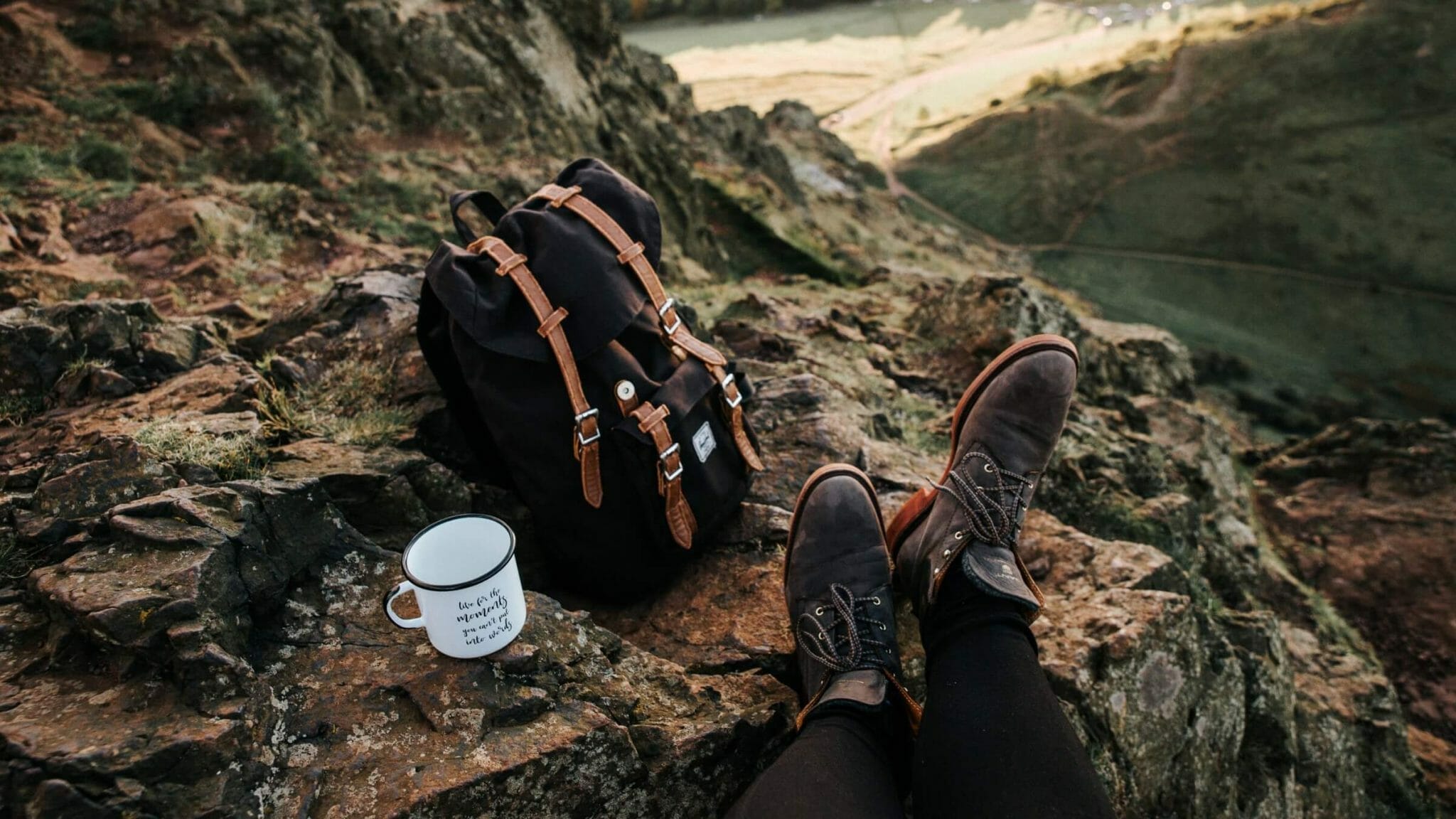 Unlaced hiking boots propped on rocks with tin camping coffee mug and strapped hiking pack.