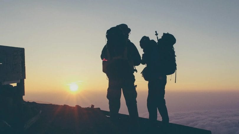 Two climbers on mountain summit at dawn with packs containing essential protective gear for hiking.