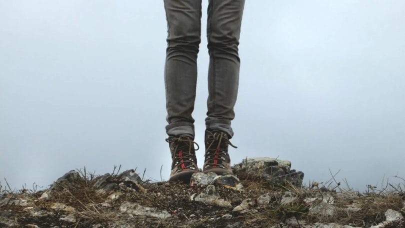 Woman with hiking boots standing on top of a rocky hill with overcast sky.